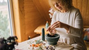 A woman in a white dress using a mortar and pestle with dried herbs on a wooden table.